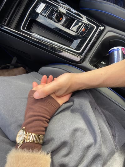 The image displays an interior view of a modern vehicle in Broxbourne, United Kingdom. Two individuals are present, their hands clasped together over gray fabric trousers. The hand on the left belongs to a person wearing a brown sleeve, possibly an arm warmer, under a lighter, fluffy beige garment. A gold-toned wrist watch with a white face is worn on this individual's left wrist. The hand on the right, displaying a light skin tone and visible arm hair, is holding the left hand.
The vehicle's dark, glossy center console is prominent in the upper portion of the frame. It features an electronic gear selector showing "P" (Park), an "ENGINE START/STOP" button, and other control buttons including one illuminated red for "AUTO HOLD" and another marked "P" for a parking brake. A storage compartment is visible below these controls. To the right of the console, a cupholder contains a silver beverage can with a pink and white label. The vehicle's seat, dark gray with blue stitching, is visible on the right. A blurred, light brown object is partially visible on the floor in the bottom left corner.