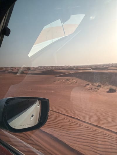 Captured from inside a vehicle, the image reveals a stark desert landscape in Al Madam, United Arab Emirates. The sandy terrain exhibits a textured pattern of ridges and furrows. A side mirror of the car is visible, reflecting a glimpse of the vehicle's interior. The windshield reflects a hazy sky and a portion of the vehicle’s dashboard.