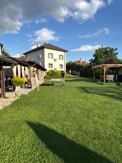 This image captures an outdoor residential setting in Hristo Danovo, Bulgaria, under a partially cloudy blue sky with the sun positioned to cast long shadows from left to right across a large green lawn, suggesting late afternoon or early evening.
On the left, a dark-wood covered patio houses a round table with dark chairs, where one shirtless male in dark shorts is seated. Standing barefoot beside him, another shirtless male in light shorts holds a green bottle and interacts with the seated individual. A small, spherical, potted green plant is situated near the patio.
In the mid-ground, a light-colored outdoor ping-pong table is positioned on the grass. To its left, a male wearing a dark long-sleeved shirt and dark bottoms stands near the table, holding a white object. Several metallic spheres, possibly bocce or petanque balls, are scattered on the lawn in this area. Further right, under a wooden pergola, are two dark-colored outdoor hot tubs or spas, one with a cover, and an integrated wooden bench.
The background is dominated by a two-story, light-colored house with a dark brown sloped roof, featuring multiple windows, air conditioning units, and two white satellite dishes mounted on its exterior. Another shirtless male in light shorts is visible walking away towards the right background. The property's perimeter is lined with various trees, shrubs, and neatly trimmed hedges, with glimpses of distant residential roofs. The foreground lawn consists of a mix of grass and clover.