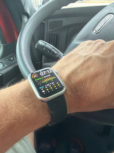 In Wildwood, United States, an individual is driving a Freightliner truck, as indicated by the logo on the steering wheel. The subject's left arm, adorned with a smartwatch displaying the time "07:12" and weather information, rests on the steering wheel. The smartwatch shows a temperature of 22 degrees Celsius and forecasts for the upcoming days, with icons depicting sunshine and varying degrees of cloudiness. The environment appears to be an interior of a vehicle, likely a truck cab, with the steering wheel and dashboard visible. The overall lighting suggests it is daytime.