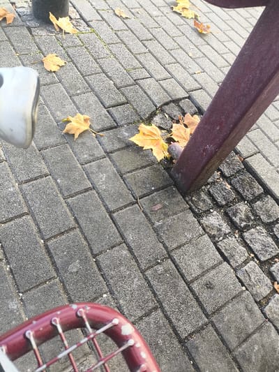 The image depicts a ground-level view in Freital, Germany, focused on a paved pedestrian surface during autumn. The primary paving consists of rectangular, light-grey concrete pavers laid in a running bond pattern, which transitions to a section of darker, irregularly shaped cobblestones on the right. Scattered across both paved areas are approximately a dozen dry, yellow-orange deciduous leaves. A weathered, dark reddish-brown rectangular beam or support structure extends diagonally from the upper right towards the lower right, appearing anchored at the interface between the two paving types. In the upper left quadrant, a portion of a light-colored, curved metallic object, consistent with a bicycle fender or fork component, is visible, partially obscuring a dark cylindrical post or pole. In the lower left quadrant, the upper section of a sports racket, characterized by a chipped reddish frame and a white string grid, is present. No human or animal subjects are depicted, and no specific actions or interactions are occurring.