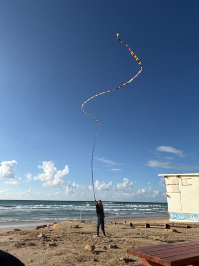 A daytime scene at Beit Yanai beach, Israel, shows a lone individual engaged in an activity on the sandy shore. The person, dressed in a dark jacket and blue jeans, stands facing the ocean, holding a long, slender, dark-colored pole that arches upwards. Attached to the top of this pole is a long, multi-colored (rainbow-patterned) streamer or kite, which extends further into the clear blue sky, displaying a gentle S-curve shape. The sky features scattered white cumulus clouds.
To the left, the Mediterranean Sea exhibits moderate waves, with several figures visible in the water further out. A pier or distant breakwater is visible on the horizon. The beach itself is sandy with some rocks and eroded sections near the waterline. To the far right of the frame, a cream-colored, single-story wooden building with boarded-up windows is situated on the sand, featuring a blue wave-like mural near its base. In the immediate foreground, the corner of a reddish-brown wooden picnic table is partially visible. The overall environment suggests an active beach setting under fair weather conditions.