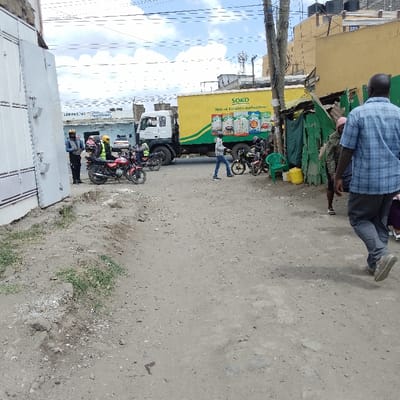 The scene depicts an unpaved, dusty road or open area in Ongata Rongai ward, Kenya, bordered by commercial structures under a partially cloudy sky.

In the foreground, the ground is light brown-grey dirt with minimal sparse vegetation on the left edge. To the far left, a tall, white corrugated metal gate or wall structure is visible.

In the mid-ground left, several motorcycles are parked or idling. Three individuals are visible near these motorcycles; two wear yellow reflective vests and helmets, one standing near a red motorcycle. A third individual in a dark uniform (blue shirt, dark pants) stands near another motorcycle further left.

Dominating the center-mid-ground is a large white truck with a bright yellow cargo container. The container is emblazoned with the green "SOKO" logo and the Swahili slogan "Mio ni kuanza mahusiano" alongside images of various consumer products such as cooking oil and flour. The truck appears to be stopped or moving slowly.

To the right of the truck, a person wearing a blue t-shirt and jeans walks from left to right, past the rear wheels of the truck, towards a cluster of motorcycles parked near a green makeshift structure. Adjacent to these parked motorcycles, a light green plastic chair and two yellow plastic jerrycans are placed.

The right side of the image features a building with a yellow facade, topped with black water tanks. Below this building, several stalls constructed from green corrugated iron are visible.

In the immediate right foreground, a male individual is seen from behind, walking towards the right. He wears a blue and white plaid shirt, dark pants, and grey shoes. Another person is partially obscured behind him.

Overhead power lines traverse the sky, and in the distant background, urban buildings with communication towers or antennas are visible. A sign for "Eurofresh" is faintly discernible on a building in the far background-left.