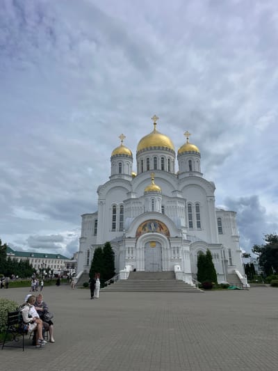 The image shows the Holy Trinity Cathedral in the city of Diveyevo, Russia, under a cloudy sky. The large, white cathedral features multiple golden domes topped with crosses. The entrance has a wide staircase leading up to ornate doors, with a mosaic above the entrance. Several people are visible in the foreground and background, including two women sitting on a bench on the left, and others walking around the plaza in front of the cathedral. To the left of the cathedral, there are other buildings with green roofs, and trees are scattered throughout the scene. The ground is paved with grey interlocking bricks.
