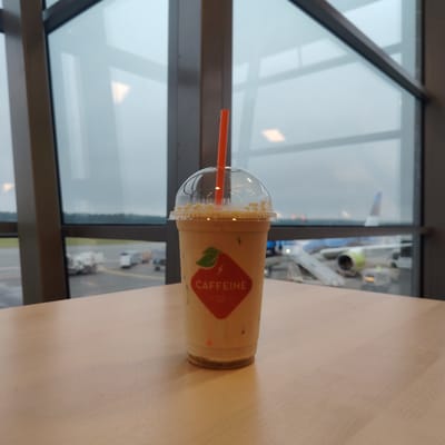The image depicts an iced beverage in a clear plastic cup with a domed lid and an orange straw, positioned on a light-toned wooden tabletop in the city of Skulte, Latvia. The cup features a red, shield-shaped label with a green leaf and lightning bolt logo, and the text "CAFFEINE EST. 2007". Through large, dark-framed windows in the background, an airport tarmac is visible. A commercial airplane, predominantly blue and white with a green-ringed engine nacelle, is parked at a gate with attached boarding stairs on the right side of the frame. Multiple ground support vehicles are present on the tarmac to the left. A distant treeline is visible under an overcast, gray sky, and the tarmac appears damp.