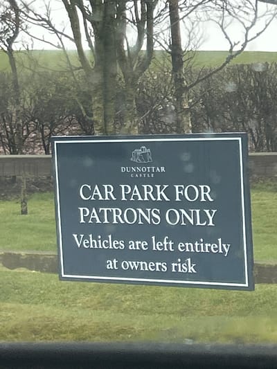 The image depicts a sign indicating the car park is for patrons only at Dunnottar Castle, located in Stonehaven, United Kingdom. The sign is dark with white lettering and a white border. Underneath "CAR PARK FOR" is the Dunnottar Castle logo. The sign also states "Vehicles are left entirely at owners risk." The sign is positioned outdoors, near what appears to be a tree without leaves and a grassy area. The image is somewhat obscured, possibly taken from inside a vehicle.