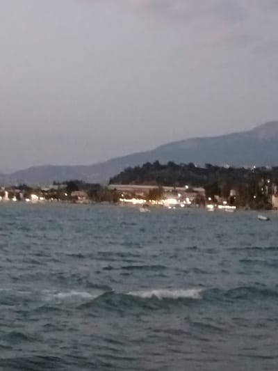The image depicts a coastal scene at dusk or dawn. In the foreground, a body of water with gentle waves is visible. The water appears dark and rippled. In the midground, a shoreline is lined with buildings and illuminated by numerous lights, suggesting a populated area. Trees and vegetation are interspersed along the coastline, and hills or mountains form a backdrop to the settlement. A few small boats are discernible on the water, one to the right and others further left. The sky is overcast with a muted light. The location appears to be a coastal town or resort area within the Municipal Unit of Vrachneika, Greece, given the natural landscape and the presence of illuminated buildings along the water.