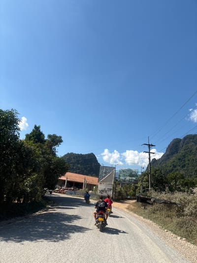 The image depicts an outdoor daytime scene in Hong Ngern Village, Laos, on a light-colored road traversing a mountainous, vegetated landscape under a clear blue sky with scattered white cumulus clouds. Three individuals are riding scooter-style motorbikes away from the viewer along the road. The closest rider, wearing a black T-shirt, operates a yellow motorbike displaying a Lao license plate "ປທ 7834". Ahead, a rider in a red T-shirt is on a yellow motorbike, followed by a third rider in a blue T-shirt on a dark-colored motorbike.

To the left of the road, dense green foliage obscures a large building with a terracotta-colored tiled roof. In front of this building, several small recreational vehicles, consistent with ATVs or buggies, are parked. Shadows from roadside trees are cast across the left side of the road. To the right of the road, a utility pole with multiple power lines is present. Further into the mid-ground on the right, two large signs flank what appears to be an entrance or gateway; the left sign is dark and obscured, while the right sign is rectangular, featuring blue and white elements with Lao script. Beyond the immediate roadside, prominent karstic limestone mountains, heavily covered in green vegetation, rise in the background on both sides of the visible path. Sparse, dry vegetation lines the immediate right edge of the road.