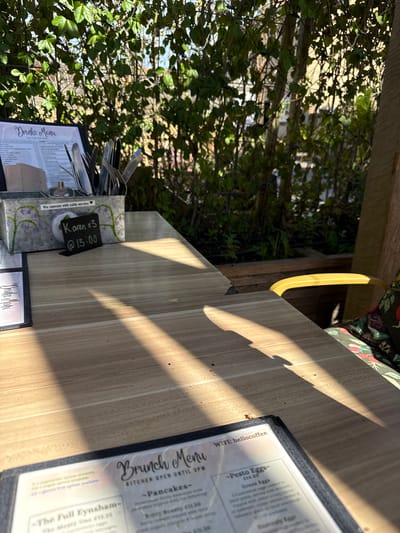 A table at a cafe or restaurant in West Oxfordshire, United Kingdom, shows a menu with "Drinks Menu - Served All Day," "Brunch Menu - Kitchen Open Until 3PM" and WIFI: hellocoffee printed. A card with "Karen x3 @13:00" indicates a reservation for three people named Karen at 13:00. Cutlery in a container, "We operate with table service❤❤❤" printed on the side of the container, is also on the table.