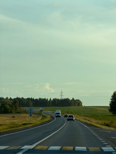 This is a photograph of a rural road with light traffic under a pale sky. The road is paved and has white lane markings. A pedestrian crossing with yellow and white stripes is visible in the foreground. Several vehicles, including a white truck and a silver car, are traveling on the road. The surrounding landscape consists of green fields and a line of trees in the background, with a power pylon visible in the distance. The location is identified as Togashevo, Russia, implying it's a rural area outside of a major city. The scene captures a moment of transit on a potentially scenic route, likely during daylight hours given the ambient light. There are no people directly visible in the frame, though their presence is implied by the vehicles. The overall atmosphere is calm and uneventful.