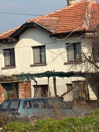 The image depicts a residential scene in Kermen, Bulgaria. A two-story building with light-colored, possibly stucco, exterior walls and a red clay tile roof occupies the central and upper portions of the frame. The roof tiles exhibit displacement and weathering. A dark-colored eaves structure with exposed wood soffit is visible beneath the roofline. Two rectangular, dark-framed windows with brown sills are prominent on the upper story facade; multiple white electrical wires are strung horizontally across this facade. Vertical cracks are visible in the stucco on the left side of the upper story.
In the lower mid-ground, a partial auxiliary structure is visible, featuring exposed red brick on the left and a weathered, dark green wooden double door. Above this, a crude overhead trellis structure, supported by wooden posts, is covered with dark green mesh material and intertwined with bare vine branches. A chain-link fence runs behind this structure.
In the lower foreground, a light blue minivan or station wagon is partially obscured by dense, dry, leafless vegetation and green ground cover. Bare tree branches extend from the right side of the frame, casting shadows across the building. The sky above is clear and light blue. No people are visible.