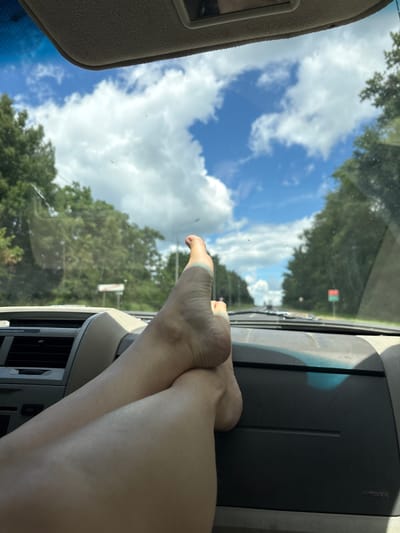 This media file depicts a close-up view of a person's legs and feet resting on the dashboard of a vehicle. The scene appears to be captured from inside the car, looking out through the windshield. The sky is a vibrant blue with scattered white clouds, suggesting a daytime setting, likely mid-morning or afternoon due to the bright sunlight. Lush green trees line the road visible in the distance, hinting at a rural or suburban environment. A traffic light is visible in the background, indicating the presence of a road and potentially traffic. The interior of the car is partially visible, including the dashboard with a textured surface and an air vent. The overall mood is relaxed and casual, with the person choosing to rest their feet on the dashboard. The location is identified as Глебовское, Russia. No discernible text is present in the image.