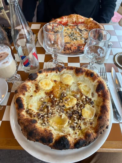 The image depicts a dining table scene at a restaurant in Deauville, France. In the foreground, a large, round pizza with a charred crust rests on a white plate. This pizza is topped with melted white cheese, several circular pieces of goat cheese, and a generous scattering of chopped walnuts, drizzled with what appears to be honey or olive oil. To the right of this pizza are a metal fork and knife resting on a white napkin.
Behind the foreground pizza, slightly out of focus, is another large, round pizza on a white plate. This second pizza features a red tomato base, anchovies, and green capers.
The table surface is a brown and cream-colored checkerboard pattern. On the left side of the table, a clear glass bottle with a label featuring red, white, and blue elements (resembling the French flag) and the word "Deauville" is visible. Next to the bottle is a clear glass mug containing a light brown, frothy beverage, possibly a latte or hot chocolate. Two empty stemmed wine glasses are positioned between the two pizzas. A silver salt shaker is visible behind one of the wine glasses on the right. In the blurred background, a person wearing a dark top is partially visible, seated opposite the viewer, and the hint of light-colored, possibly wicker, furniture is present. The scene captures a meal in progress.