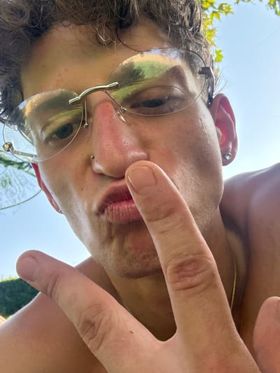 A close-up, overhead shot shows a person's face from the nose up, against a bright blue sky. The person is wearing round, transparent-rimmed glasses that reflect the surroundings. They have curly brown hair and a nose ring. Their mouth is puckered, and a finger is placed just above their lips. Another finger is raised in a peace sign, and a portion of their shoulder is visible, suggesting they are shirtless. The environment appears to be outdoors, with hints of green foliage in the background, consistent with the rural setting of Villa del Prado, Spain, under a clear sky.