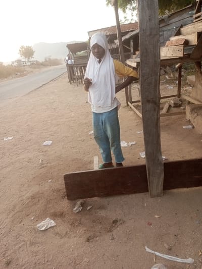 A girl in a white headscarf, yellow shirt, and blue pants stands outdoors. She has one hand on her hip and faces forward. She is near a wooden structure with tables, possibly a market stall, and a large wooden post. The ground is dirt with scattered trash. A road is visible in the background along with other people and buildings.