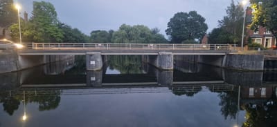 The image depicts a concrete bridge spanning a calm body of water, likely a canal or river, in the city of South Holland, United Kingdom. The time appears to be dusk or early evening, as evidenced by the dim lighting and the streetlights casting a glow.

The bridge itself is a utilitarian structure with concrete abutments and a flat deck. A pale railing runs along its length, and a vehicle's headlights are visible as a blur on the left side of the bridge, indicating it is in use. The water below acts as a mirror, reflecting the bridge, the surrounding trees, and the sky with remarkable clarity.

On the banks of the water, a variety of trees are visible, ranging from large, mature deciduous trees to weeping willows. On the right bank, a brick building with white-framed windows can be seen, partially obscured by foliage and illuminated by a streetlight. The overall atmosphere is tranquil, with the stillness of the water enhancing the serene mood.