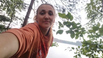 A woman with light brown hair pulled back is taking a selfie outdoors. She is wearing an orange hooded sweatshirt. She is positioned in the foreground, with trees and foliage framing her. Behind her, a body of water is visible, with a treeline on the opposite shore. The overall lighting suggests an overcast day. The location is identified as Tarasovskoe, Russia.
