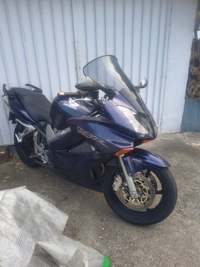 The image shows a dark blue Honda VFR motorcycle parked outdoors on a concrete surface. The motorcycle is a sport-touring bike with a prominent windshield and fairings. It has a black seat, silver accents, and gold-colored brake discs. The front wheel is visible, as is part of the engine and exhaust system. A "Honda" logo is visible on the side of the motorcycle. The background consists of a light blue, ribbed wall and a concrete ground with some scattered leaves. The lighting suggests it is daytime, with the sun casting some shadows. There is no activity taking place; the motorcycle is stationary. The scene appears to be in a residential or storage area. No people are visible in the image. There is also a roll of white, mesh-like material visible in the lower right corner of the image.