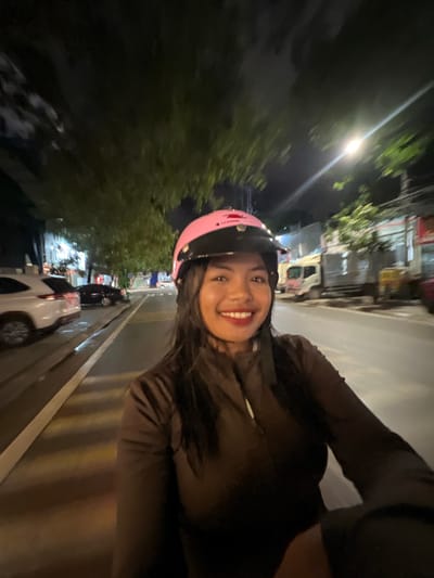 A woman is on a motorbike at night, wearing a pink helmet and a dark brown jacket. She is smiling and looking at the camera. The street is wet, suggesting it has rained recently. There are trees, parked cars, and a truck visible in the background. The scene appears to be in a city. Based on visual cues such as vehicle types and road markings, this scene is consistent with a location in Minh Xuân Ward, Vietnam.