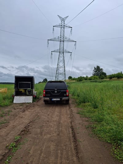The image shows a rural scene in Lubnica, Poland, dominated by a large electricity pylon and a dirt track. A black Nissan Navara pickup truck is parked on the track, facing away from the camera. To the left of the truck is a black utility trailer with its rear door open, revealing some equipment inside. The surrounding environment consists of a green field with tall grass and scattered trees in the background, under a cloudy sky. The ground of the track is muddy with tire tracks visible. A yellow safety fence is partially visible on the left side of the frame.