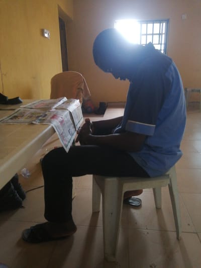 A person sits at a table with newspapers spread out on it. The person is wearing a blue shirt and black pants, and is looking down at the newspapers. The scene is indoors, with yellow walls and a tiled floor. A window with bars is visible in the background, letting in bright sunlight. There are also some chairs and other objects in the room. The overall impression is of a casual indoor setting, possibly a waiting area or a common room. The time of day appears to be daytime due to the bright sunlight. The location context is Ajasse Ipo, Nigeria.