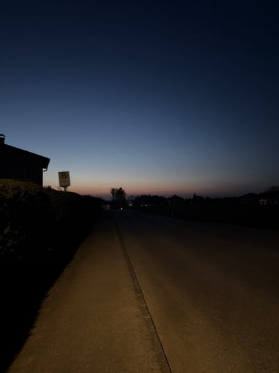 The image displays a deserted street scene in Ferschnitz, Austria, captured at late dusk or night. The sky presents a gradient from deep navy blue overhead to lighter blue, transitioning to a narrow band of orange and pink along the horizon. A faint, thin white line, consistent with an aircraft contrail, is visible in the upper sky.

In the left foreground, a paved sidewalk runs parallel to a two-lane asphalt road, separated by a curb. A dark, dense hedge extends along the left side of the frame, partially obscuring the lower section of a silhouetted building positioned further back. Above the hedge, a white rectangular sign featuring a yellow circular inset with a black 'H' (indicating a bus stop) is clearly visible.

The asphalt road extends into the distance, appearing dark but with subtle illumination from an unseen source. Silhouettes of trees and additional distant buildings line both sides of the road, particularly in the midground and background. Several faint points of light, consistent with distant streetlights or vehicle headlamps, are discernible further down the road. No individuals, vehicles, or specific actions are present within the frame.