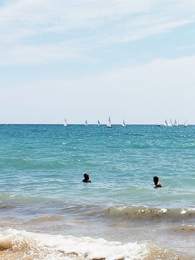The image depicts a bright, sunny day at a beach with a clear blue sky and scattered white clouds. The ocean is a vibrant turquoise, with gentle waves lapping at the shore. Several sailboats are visible in the distance, dotting the horizon. In the foreground, two people are swimming in the ocean, their heads and shoulders visible above the water. The person on the left is looking towards the right, while the person on the right is looking towards the sky. The water is choppy in some areas, with white foam from the waves. The beach itself is visible in the lower portion of the image, with sand and some seaweed. The overall mood is one of leisure and enjoyment on a warm summer day. The location is Rota, Spain, as indicated by the context.