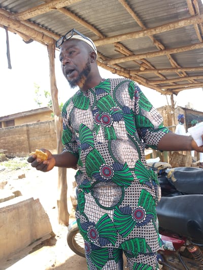 The image shows a man standing under a makeshift shelter in Iseyin, Nigeria. He is wearing a vibrantly patterned green and white African print outfit and a white cap, with glasses perched on top. He has a beard and appears to be speaking animatedly, with his mouth open. In his left hand, he holds a piece of what looks like fried food.

The shelter is constructed with corrugated metal roofing supported by rough wooden beams and poles. Sunlight streams through the gaps in the shelter, indicating it is daytime, likely a sunny day. The background reveals a dusty, rural environment with a brick wall and a building. Two other figures, one in a white garment and blue cap, and another person further in the background, are visible. A motorcycle with black seats is parked to the right of the main subject, with visible suspension and a red fuel tank. On the motorcycle, a sticker with text is partially visible, reading "BM 100" and other unreadable lines. The overall scene suggests an outdoor gathering or marketplace in a Nigerian town. The man's expression and posture convey engagement and animation, possibly in conversation or giving a speech.