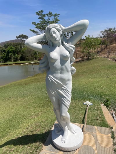 A white, unpainted concrete statue of a nude female figure stands outdoors under a clear blue sky in Pirenópolis, Brazil. The statue depicts a woman with her arms raised, hands positioned behind her head, and a drapery covering her lower body from the hips down. Her chest is exposed. The statue is anchored on a round, flat concrete base which rests on a paved area composed of brown and grey geometric tiles. To the left of the statue, a manicured green lawn extends to a body of water, likely a pond or lake, with its far bank lined by lush green trees. In the background, rolling, tree-covered hills are visible. To the right of the statue, the terrain appears drier, with sparse vegetation and a visible stick protruding from the grass, topped with a small, light-colored object, possibly a piece of fabric or a discarded sandal. The scene is illuminated by strong sunlight, casting a distinct shadow of the statue to its left.