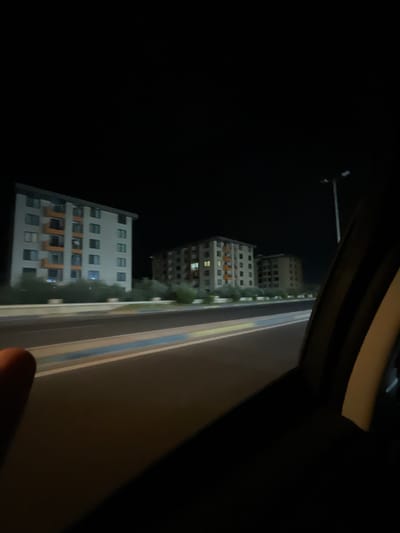 The image shows a night scene in the city of Sumgayit, Azerbaijan, viewed from inside a moving vehicle. The foreground is dominated by the dark interior of the car, with a portion of the passenger-side window frame visible. In the middle ground, a road with distinct white and blue lane markings streaks by. Beyond the road, two apartment buildings are visible. The building on the left is taller and appears to be a light color with orange balconies. The building on the right is shorter and has visible windows lit from within. The sky is completely dark, indicating it is nighttime. Scattered green foliage can be seen between the road and the buildings. A street lamp is also visible in the distance on the right side of the frame. The overall impression is of a nighttime drive through a residential area.