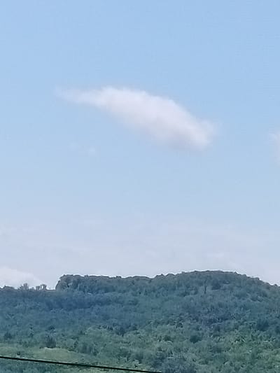 A landscape scene features a distant, undulating, tree-covered hill against a pale blue sky with a single, wispy cloud. The hill's summit is uneven and rocky. In the foreground, two dark, horizontal lines, likely power lines, stretch across the bottom of the frame. The overall impression is one of a rural or undeveloped area, possibly in the vicinity of Chișcăreni, Moldova, given the context.