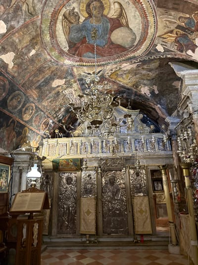 The image shows the interior of a church, likely in Kassopaia Municipal Unit, Greece, featuring ornate religious iconography and architectural elements. The ceiling is adorned with faded frescoes depicting religious figures, including an angel at the apex. A large, multi-tiered brass chandelier hangs prominently in the center of the frame. Below the ceiling, a decorative iconostasis is visible, featuring embossed silver icons of saints and religious scenes, interspersed with hanging votive lamps. In the foreground, a wooden lectern holds an open book, and to the left, a framed icon is displayed. The floor is tiled in a diamond pattern of red and beige squares. The overall impression is one of historical and religious significance, with aged artwork and traditional church furnishings.