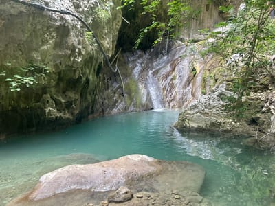 A serene natural landscape within the Municipal Unit of Ellomenos, Greece, features a clear, turquoise pool of water nestled at the base of a rocky cliff face. A small waterfall cascades down the damp, textured rock, feeding into the pool. Lush green foliage drapes from the cliff edges and branches overhang the water. Large, smooth boulders rest partially submerged in the foreground, with smaller stones visible along the pool's edge. The overall scene conveys a sense of tranquil natural beauty.