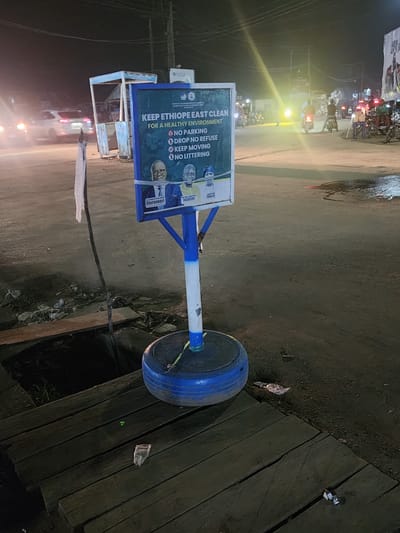 At night in Obiaruku, Nigeria, a sign promoting cleanliness in Ethiope East stands near a roadside drain. The sign, partially obscured by the angle, displays the message "KEEP ETHIOPE EAST CLEAN FOR A HEALTHY ENVIRONMENT" along with instructions prohibiting parking, dropping refuse, and littering. Images of individuals named Oborevwori, Ogedegre, and Charles are also visible on the sign. The sign is attached to a pole supported by a blue-painted tire acting as a base. The immediate environment consists of a paved road and wooden planks covering a drain. Debris and litter are scattered around the drain. In the background, streetlights illuminate a row of parked motorcycles and other vehicles, indicating a moderately busy street scene.