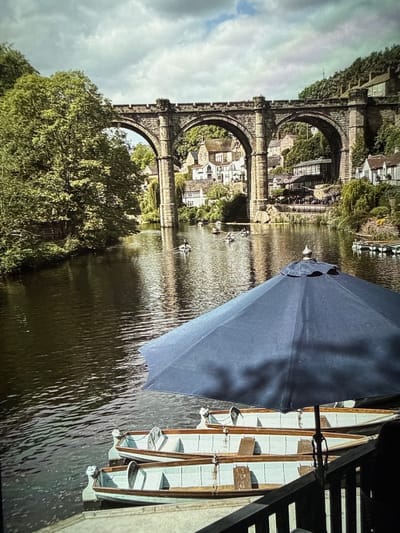 The image depicts the River Dove with a view of the Conisbrough Viaduct in Conisbrough, Barnsley, United Kingdom. The stone-arch railway viaduct is prominently featured, with trees and foliage at its base and visible buildings within its arches. A body of water, likely the river, fills much of the frame. Rowboats are positioned on the water. An open dark-colored umbrella sits on the right side, obscuring some of the rowboats. A dark railing runs along the right edge.
