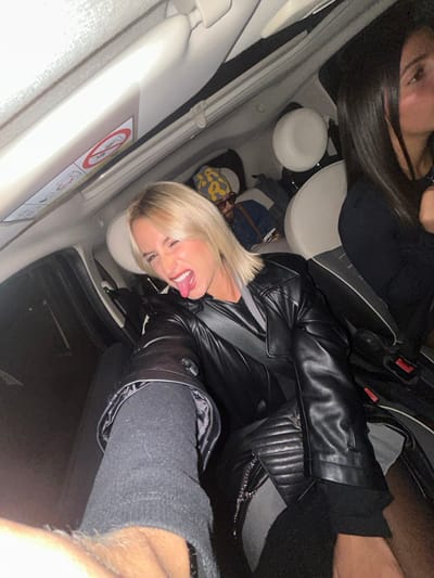 Inside a car, two women are visible. One has long, dark hair and wears a black top. The other, closer to the camera, has short blonde hair and a black leather jacket, sticking her tongue out playfully. Both appear to be wearing seatbelts. The car's interior is light-colored, with visible seats, seatbelts, and a "no smoking" sign on the sun visor.
