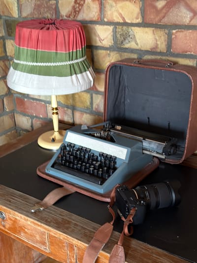 The image depicts a still life arrangement on a desk, set against a brick wall. The objects include an antique typewriter, a vintage suitcase, a camera, and a lamp with a multi-colored lampshade. The typewriter is a light blue model with black keys, resting on a brown tray. The suitcase is open, revealing a dark interior, and appears to be made of brown leather. A modern digital camera with a prominent lens and a brown strap is placed to the right of the typewriter. The lamp has a base with decorative gold accents and a lampshade featuring horizontal bands of red, green, and white fabric. The desk surface is black, and the background is a brick wall with varying shades of red and yellow. There are no people or animals present in the image. The scene is static and arranged, suggesting a curated display. The location is identified as Shabdan, Kyrgyzstan.