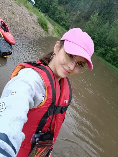 A young woman is taking a selfie by a body of water, likely a river or lake, near a wooded area. She is wearing a red life vest over a light gray hoodie and a bright pink baseball cap. She also has on round, gold-rimmed glasses and hoop earrings. On the left side of the frame, a red kayak is visible, resting on the pebbly shore next to a black waterproof bag. The water appears murky, and the surrounding landscape is lush with green trees, suggesting a natural, outdoor setting. The sky is overcast, indicating a cloudy day. The woman's expression is a slight smile, and she appears to be relaxed and enjoying herself. The overall mood is casual and adventurous. The image seems to have been taken during the daytime.