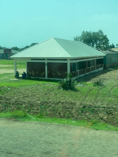 The image depicts a long, single-story building with a distinctive white, pitched metal roof. The building's exterior walls appear to be constructed of reddish-brown brick, interspersed with white vertical supports and a white horizontal band along the bottom. Numerous dark-colored windows, possibly shutters, are visible along the side of the building. Two individuals are seated on the ground in the shade of the building's overhang, one wearing a light-colored shirt and the other a dark blue garment. They appear to be interacting with each other. The building is situated in a landscape with patches of green grass and tilled soil, suggesting an agricultural or semi-rural setting within the city of Jama'a, Nigeria. In the background, other buildings and trees are visible, with a bright, clear sky overhead. The overall impression is one of a functional, possibly community-oriented structure in a developing area.
