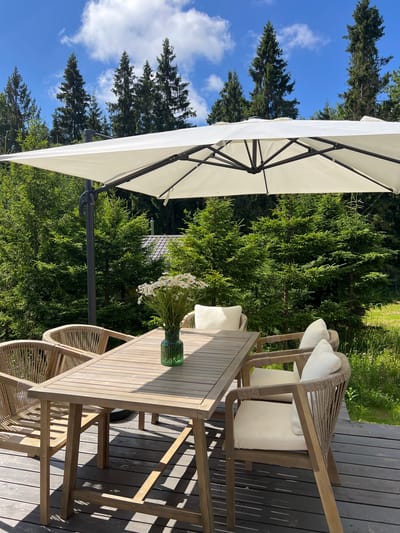 An outdoor dining set is situated on a wooden deck, surrounded by a dense forest of evergreen trees under a clear blue sky with scattered white clouds. A large white cantilever umbrella shades the table and chairs. The table is made of light-colored wood, and a vase containing a bouquet of white daisies is placed in the center. Four chairs with light-colored cushions are arranged around the table. The chairs have wooden frames and woven rope details on the backs. The deck is constructed of dark gray wooden planks. In the background, the tops of pine trees are visible, and a hint of a roofline can be seen peeking through the foliage, suggesting a nearby building. The location is Popovka, Russia.