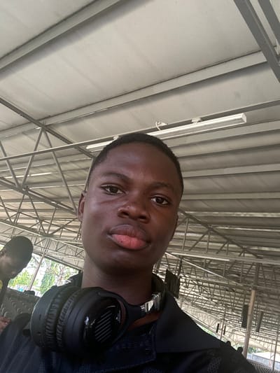 The image depicts a close-up, upward-angled view of a young male individual in Ozuaha, Nigeria. The primary subject, with a dark complexion and short dark hair, is centered in the frame, looking directly at the camera with a neutral expression. He has full lips and a small visible mark on his right cheek area. Around his neck, resting on his shoulders, are black over-ear headphones with silver accents. He is wearing a dark, collared shirt with a subtle textured pattern. In the lower-left background, the top of a second individual's head and shoulder are partially visible; this person also has a dark complexion and short dark hair and appears to be looking downwards. The environment suggests an indoor or semi-outdoor covered space, characterized by a light-colored ceiling with visible metal structural beams and trusses. Multiple linear fluorescent light fixtures are attached to the overhead structure. Beyond the immediate structure, green foliage is visible, and several dark, rectangular objects are suspended from the ceiling in the mid-ground.