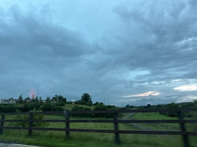 The image depicts a rural landscape under a cloudy sky, likely taken from a moving vehicle in Rathvilly, Ireland. In the foreground, a dark wooden fence with three horizontal rails runs across the frame. Beyond the fence, a vibrant green field is visible, with a gravel path winding through it towards the background. The middle ground features a gentle hillside dotted with trees and several houses, appearing to be part of a small community. A notable feature on the left side of the hillside is a bright red, almost neon, reflection or light source emanating from behind a cluster of trees. The sky is overcast with shades of gray and blue, with some hints of lighter clouds or distant sunlight breaking through on the right side of the frame. The overall impression is one of a serene, overcast day in the Irish countryside.