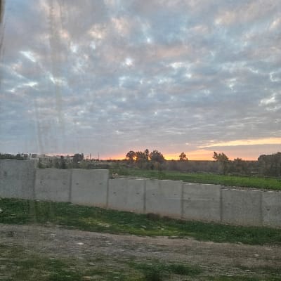 The scene captures an outdoor landscape in Ben Guerir, Morocco, at either sunrise or sunset. The sky is overcast with a dense layer of altocumulus or stratocumulus clouds, displaying grey tones with visible orange and pink coloration on the horizon, indicating the sun's position. The sun itself is a bright orange disc, partially obscured by a cluster of dark trees. In the midground, a continuous series of light grey concrete walls, likely retaining structures or barriers, extends horizontally across the frame. These walls exhibit vertical seams and regularly spaced circular marks. The ground immediately surrounding these walls consists of green grass interspersed with exposed soil. The immediate foreground is an unpaved area of dirt and gravel with sparse green vegetation. In the distant background, to the left, several low-rise buildings and construction-like structures are visible, indicating an urban or semi-urban environment. No individuals or specific actions are discernible.