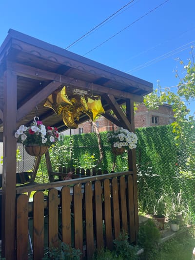 A ground-level, medium shot depicts a wooden gazebo-like structure with hanging baskets of flowers (white, red, and pink petunias), and several gold star-shaped balloons affixed to the structure's support beams. A wooden fence runs to the right, obscuring the view beyond it. In the background, beyond the gazebo, is a green wall of foliage and the upper stories of a brick building. Above the structure, clear blue sky and foliage from a vine are visible. The location is Kraishte, Bulgaria. There are no people visible in the frame, though there is a suggestion of a person to the right of the frame.