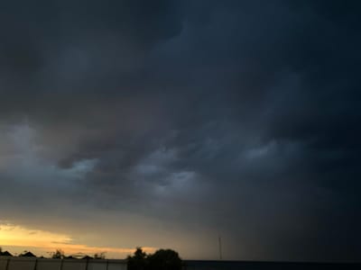 The image shows a dramatic sky with dark, heavy storm clouds dominating the upper portion. Towards the horizon, a sliver of pale yellow and orange light suggests either dawn or dusk, with some lighter clouds scattered in this area. Below the sky, a dark silhouette of trees and a fence runs across the lower edge of the frame. A faint, vertical line, possibly a utility pole or antenna, can be seen against the stormy sky in the distance. The overall scene evokes a sense of impending weather. The location is specified as Izberbash, Russia.