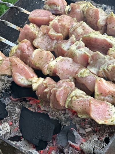 The image shows a close-up view of marinated meat skewers being grilled over hot coals. The meat is cut into bite-sized pieces and seasoned with herbs and spices. The skewers are placed on a metal grill rack, and the glowing red coals underneath indicate that the barbecue is active. The background is slightly blurred, but it appears to be an outdoor setting with greenery visible, suggesting a backyard or a picnic area. The specific location in the city of Подмой, Russia is not discernible from the image. The focus is entirely on the food being prepared.