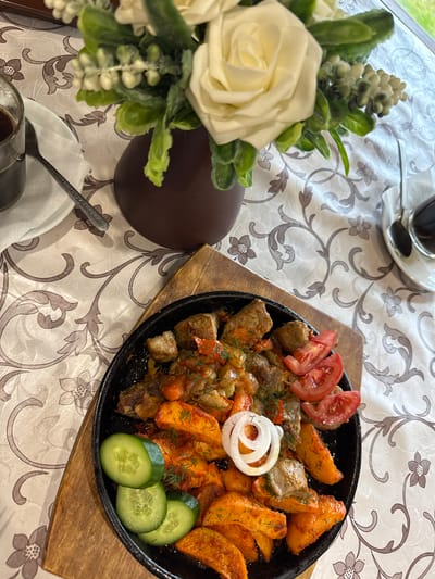 A close-up, overhead shot shows a hearty meal served on a black cast-iron skillet, resting on a wooden board. The skillet is filled with a generous portion of roasted meat cubes, intermingled with chunks of roasted potatoes and what appears to be sautéed vegetables like tomatoes and onions. Fresh dill is sprinkled over the dish, and three slices of tomato are arranged on top. Three round slices of cucumber are placed along the edge of the skillet. A single, white onion ring sits prominently in the center of the dish.

The meal is set on a table covered with a light-colored tablecloth featuring an ornate, grey floral and swirling pattern. To the left of the skillet, a bouquet of artificial white roses and green foliage is arranged in a dark brown vase, partially obscuring the lower left corner of the image. In the background, a cup of dark liquid, likely coffee or tea, sits on a saucer with a spoon. The lighting suggests it is daytime.

The overall impression is one of a satisfying and rustic meal, perhaps a home-cooked dish or a comforting offering from a restaurant. The focus is entirely on the food and its presentation, with the surrounding elements providing context for a dining scene. There is no visible text in the image, except for what appears to be embossed lettering on a spoon visible near the bottom left, which is too blurry to read. The location context of Bolotnitsa, Russia, suggests this might be a traditional Russian dish.
