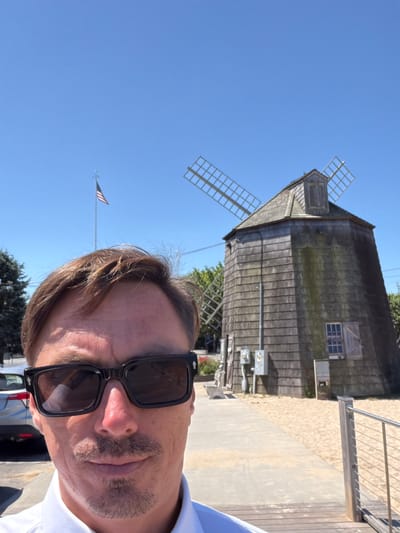 A man wearing sunglasses and a white shirt is taking a selfie in front of a wooden windmill. The windmill has four blades, two of which are visible and are made of a light-colored material. An American flag is flying on a flagpole to the left of the windmill. The ground is covered in sand, and a paved walkway leads to the windmill. In the background, there is a silver car parked to the left of the frame, and some trees and greenery. The sky is clear and blue. The scene appears to be outdoors on a sunny day in the Village of Sag Harbor, United States of America.