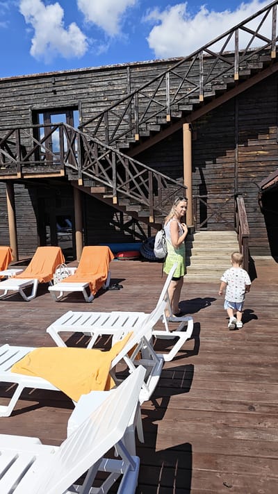 A woman with blonde hair, wearing a white top and a light green dress, stands on a wooden deck, looking to her right. She carries a white backpack on her left shoulder. A young boy, wearing a white t-shirt with a pattern and blue shorts, walks away from the camera towards the wooden stairs of a rustic, dark wood building. The building has an outdoor staircase with a criss-cross railing and appears to be an outdoor establishment, possibly a resort or cafe, given the presence of several white lounge chairs with orange and yellow towels on a wooden deck. The deck is illuminated by bright sunlight, casting long shadows. The sky above is blue with scattered white clouds. The scene suggests a warm, sunny day in a recreational area.
