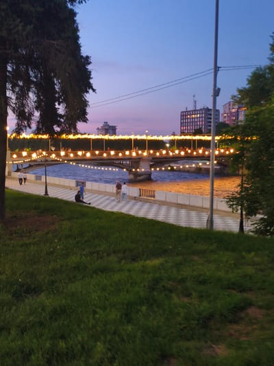 The image captures a twilight scene in the city of Atabe, Georgia, featuring a riverfront promenade and a bridge adorned with festive lights. A broad, calm river occupies the middle ground, its surface reflecting the warm glow of the lights. The promenade, paved with a checkered pattern of light and dark tiles, runs alongside the river. Several individuals are present on the promenade; two women walk together near the left side, while a man in a blue shirt stands further down. A person in dark clothing sits on the grassy bank in the foreground, facing away from the river.
The bridge, a prominent structure spanning the river, is illuminated by a continuous string of yellow lights, creating a festive ambiance. On the bridge, figures of people can be seen walking and gathered. In the background, modern buildings, including a multi-story structure with illuminated windows, rise against the deepening twilight sky. The sky itself displays a gradient of colors, transitioning from a soft pink near the horizon to a deep blue overhead, hinting at the end of day. A tall lamppost stands near the center of the frame, and lush green grass covers the foreground, adding to the park-like atmosphere. A large tree with dark foliage is situated on the left side of the image, partially obscuring the view of the sky.